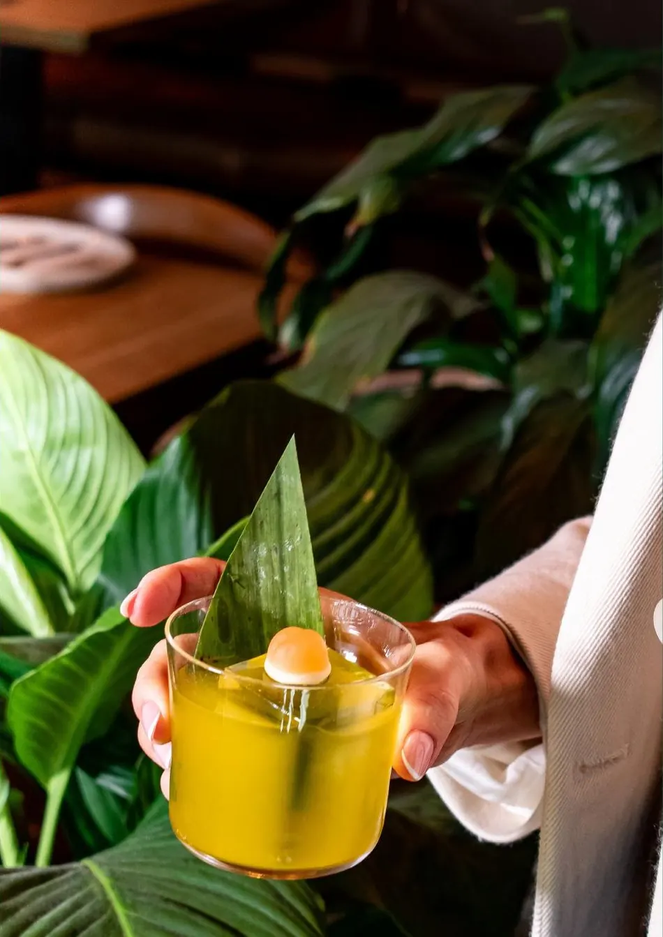 A yellow cocktail garnished with a leaf, held up against tropical plants.