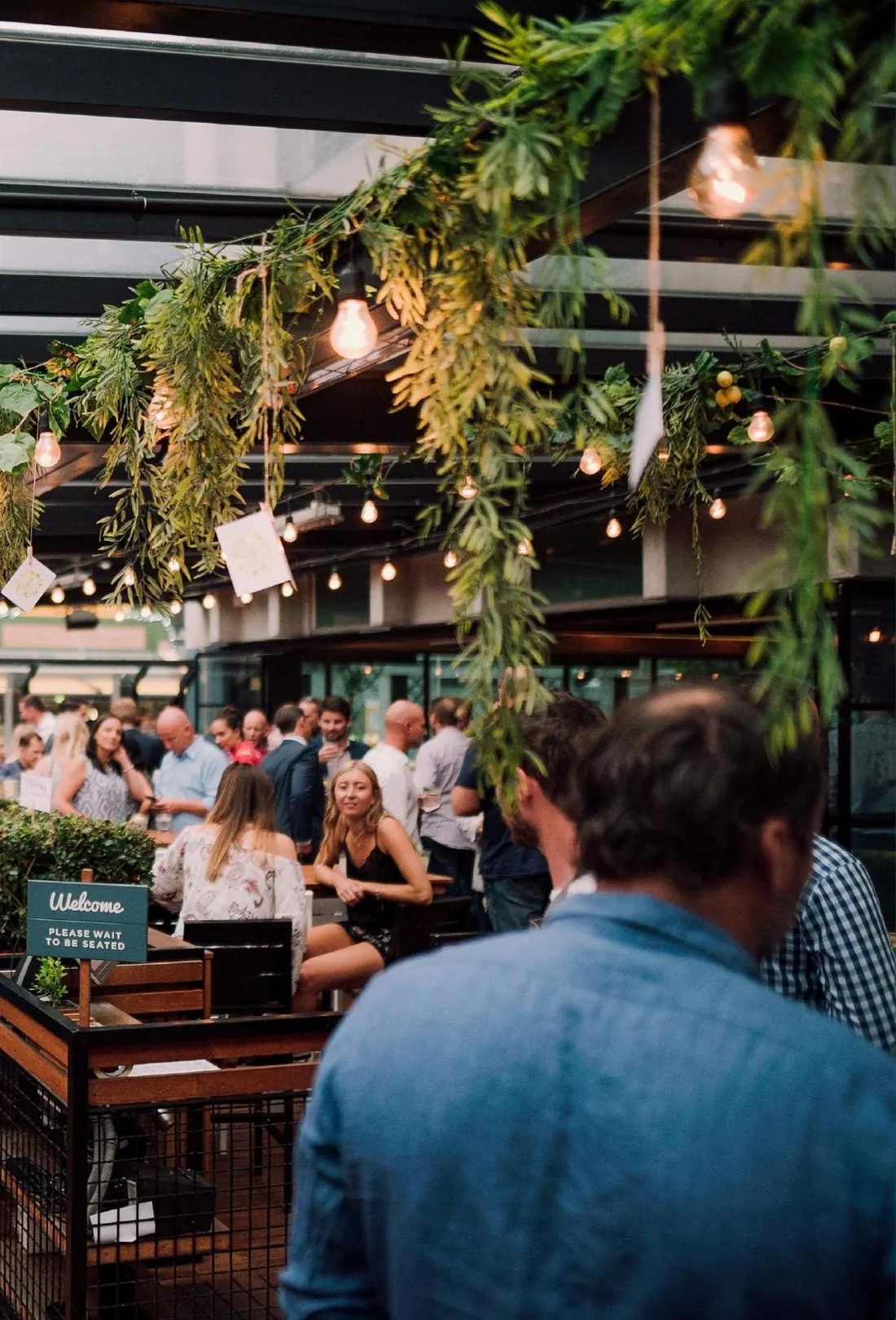 A full deck at The Glass Goose, under hanging greenery and festoon lights.